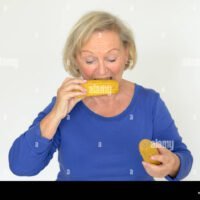 anciana-disfrutando-de-la-mazorca-de-maiz-dulce-invadiendo-los-granos-con-una-feliz-sonrisa-en-una-dieta-saludable-concepto-sobre-gris-f6xx1y